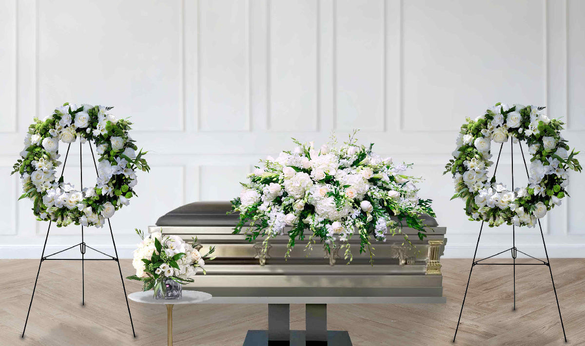 A silver casket decorated with a large white and green floral arrangement, flanked by two matching floral wreaths on black metal stands in a bright white room with wood flooring. a flower arrangement on a white and gold table placed in front of the casket