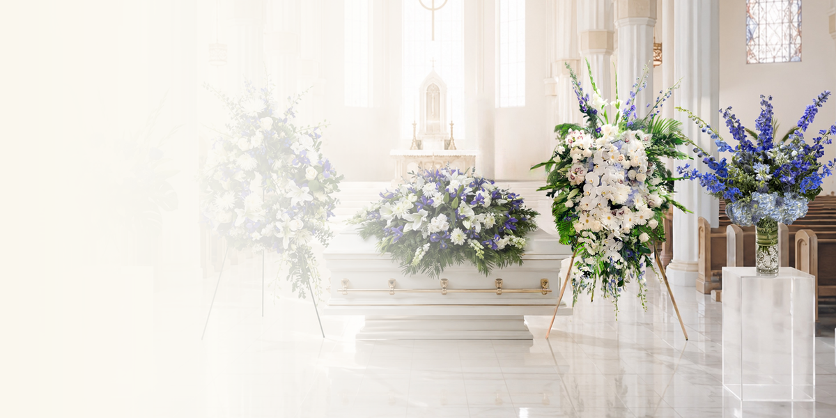 A white casket in a bright chapel or funeral hall, surrounded by large floral arrangements in white, blue, and green on stands and a clear pedestal.