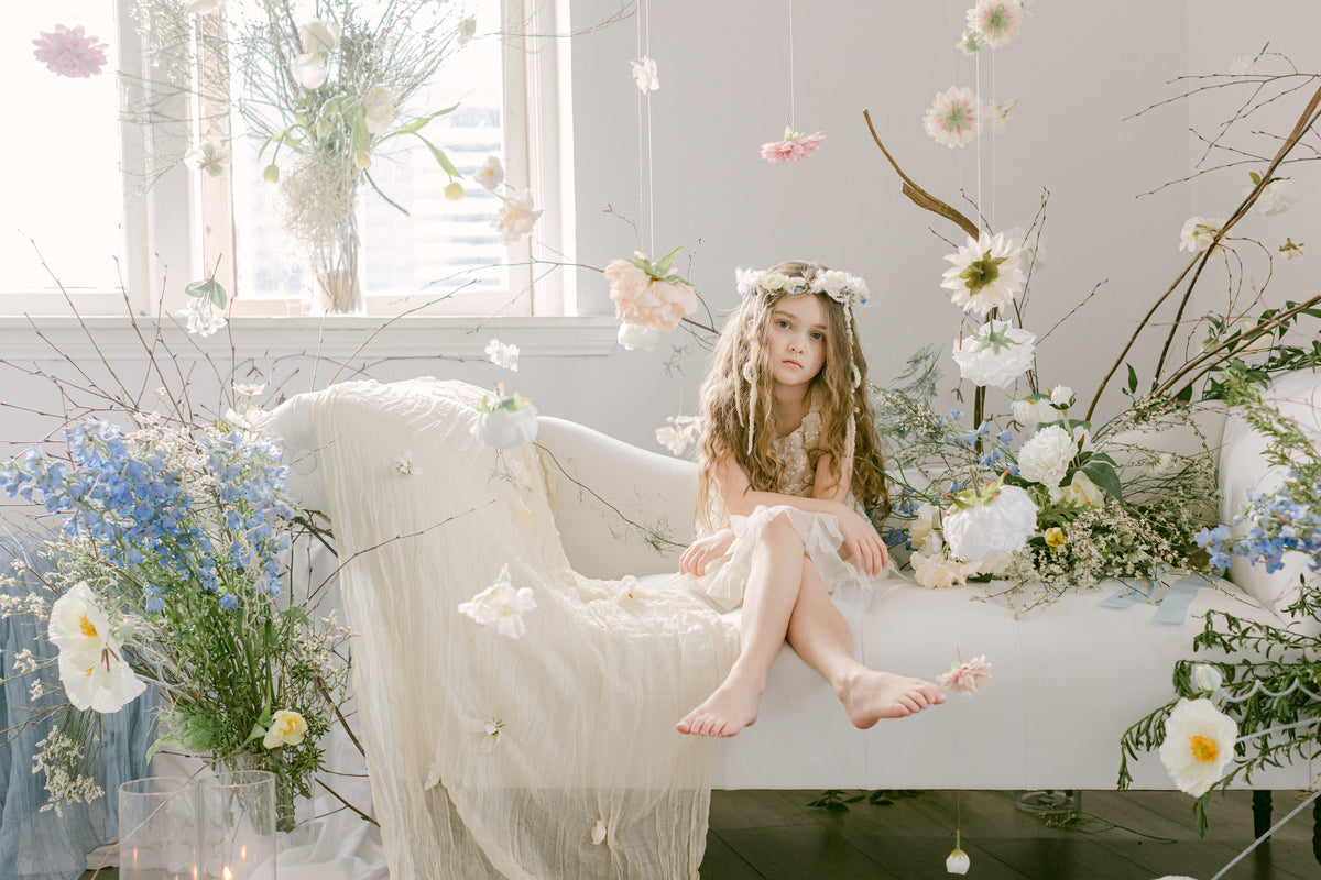 A young girl with long light brown hair sits barefoot on a white upholstered bench, wearing a white dress and a flower crown in a bright, floral-filled room. Hanging flowers, large flower arrangements, and soft natural light create a dreamy, editorial atmosphere.