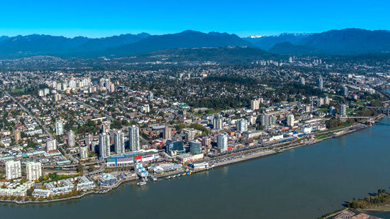Aerial view of a coastal city with dense buildings, a large river in the foreground, and mountains in the background under a clear blue sky.