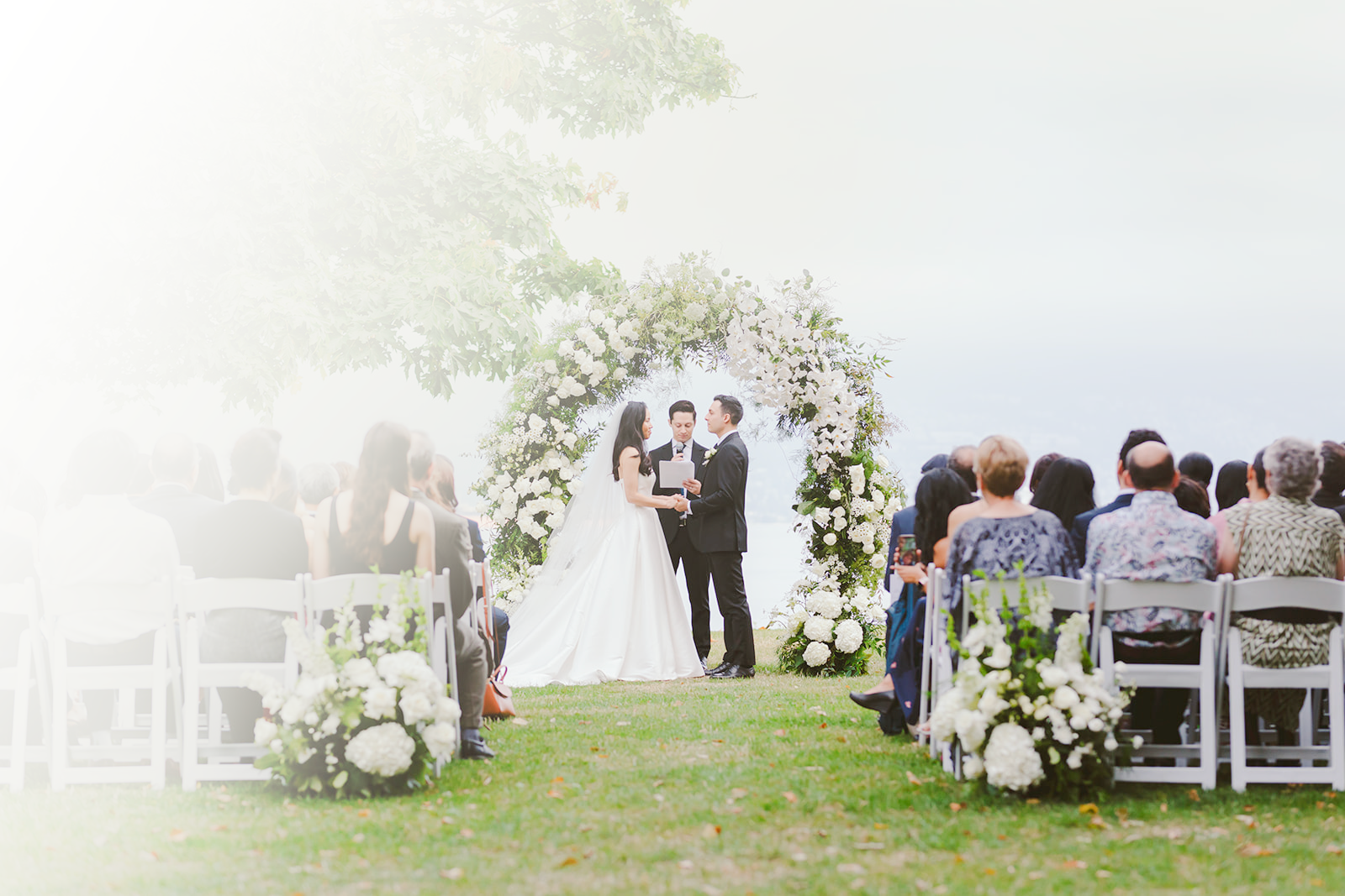 A wedding ceremony outdoors with a bride and groom standing under a floral arch, seated guests on both sides, and bright natural light with some haze across the scene.