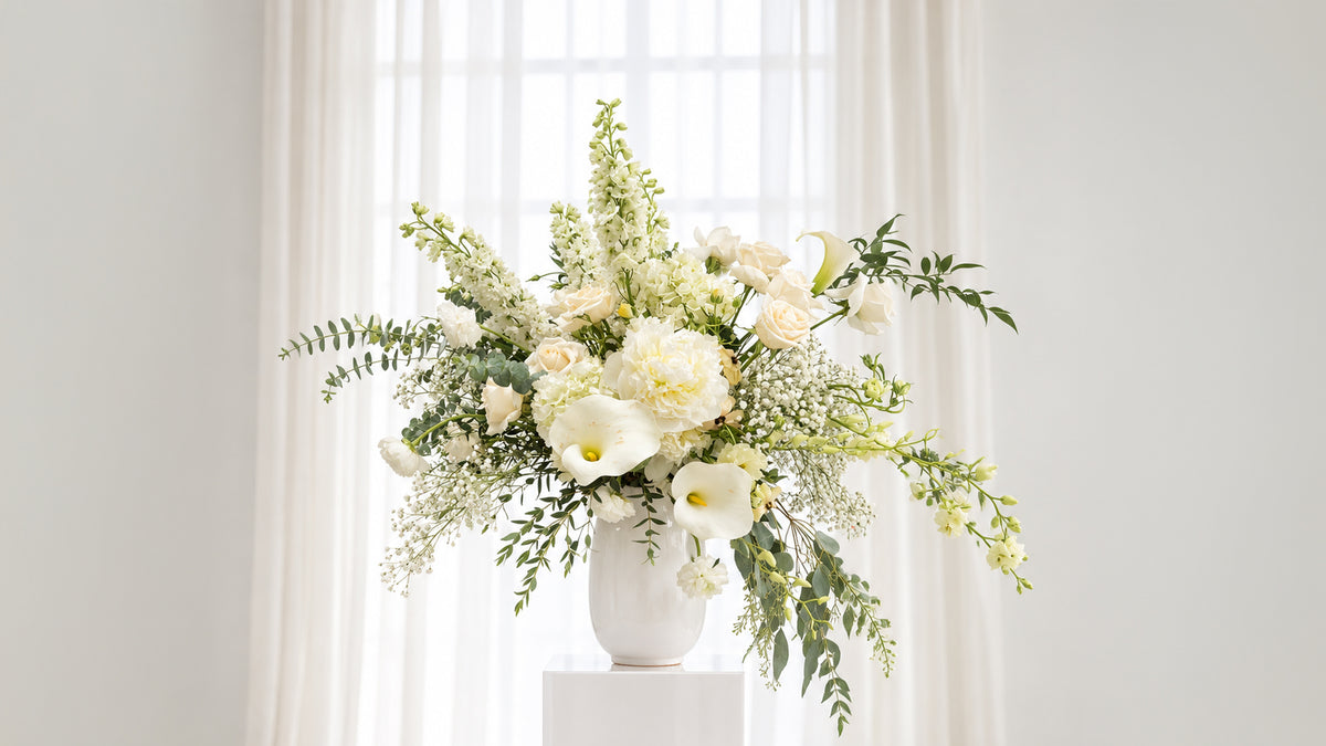 A white ceramic vase filled with a lush floral arrangement of white calla lilies, cream roses, and green foliage, displayed on a white pedestal in front of a bright window with sheer curtains.