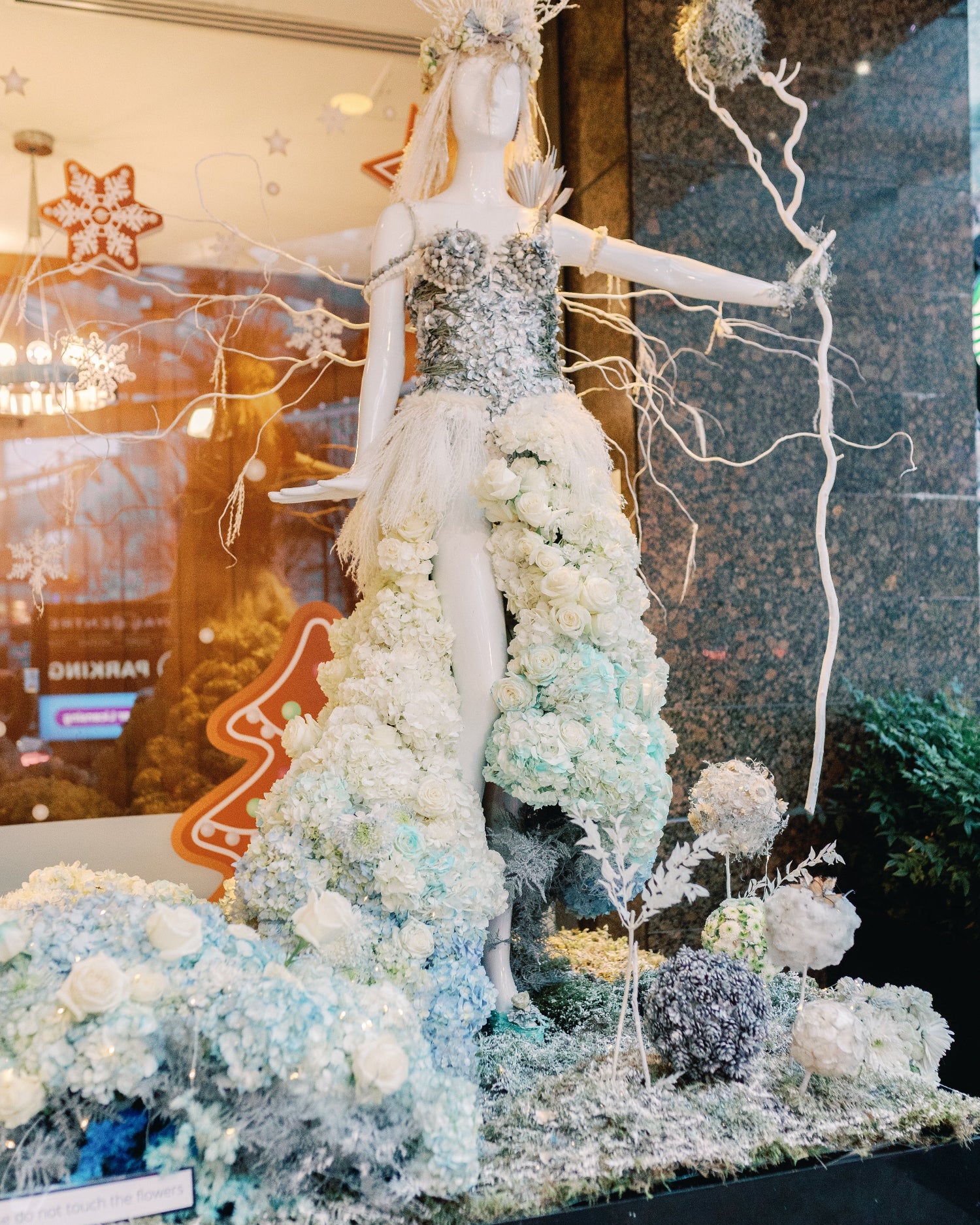 A white mannequin dressed in an elaborate fantasy winter gown made of white and pale blue flowers, glitter, and tulle, posed in a decorative display with branches and snowy floral props.