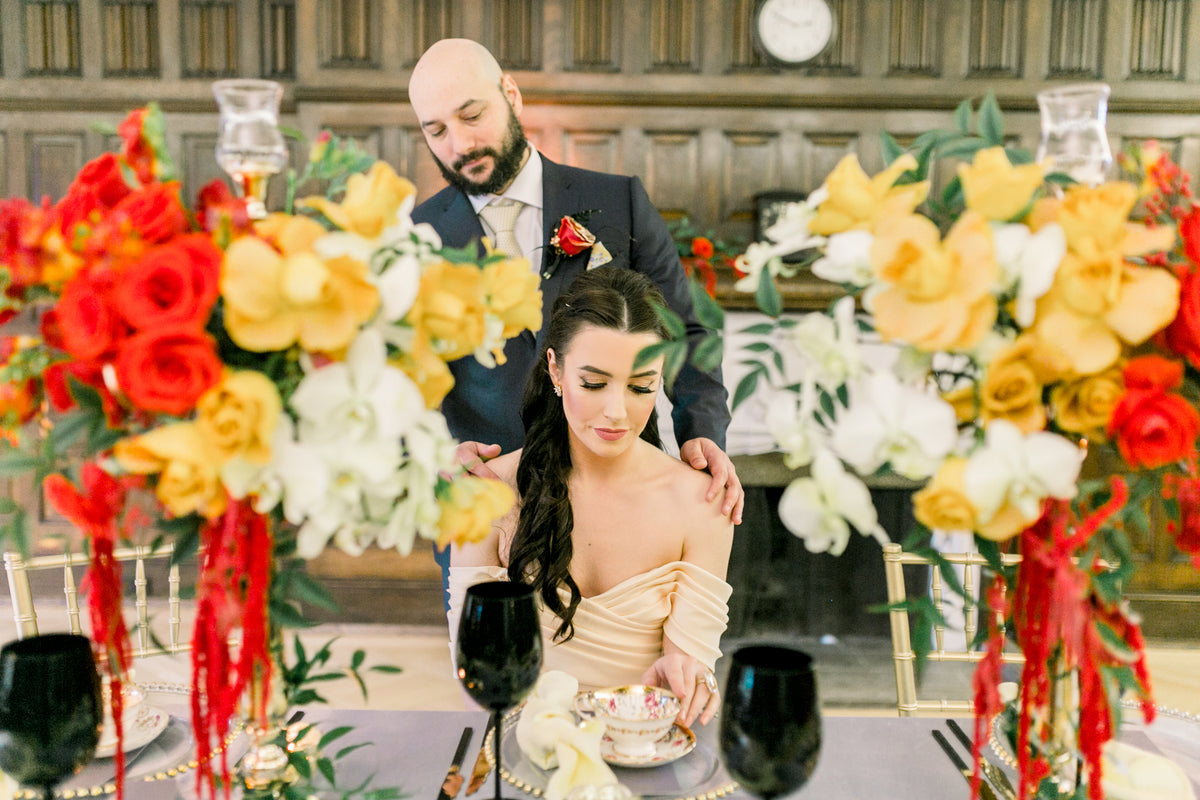 A bride in a champagne strapless gown sits at a wedding table while a groom in a dark suit stands behind her, framed by large floral arrangements in red, yellow, and white.