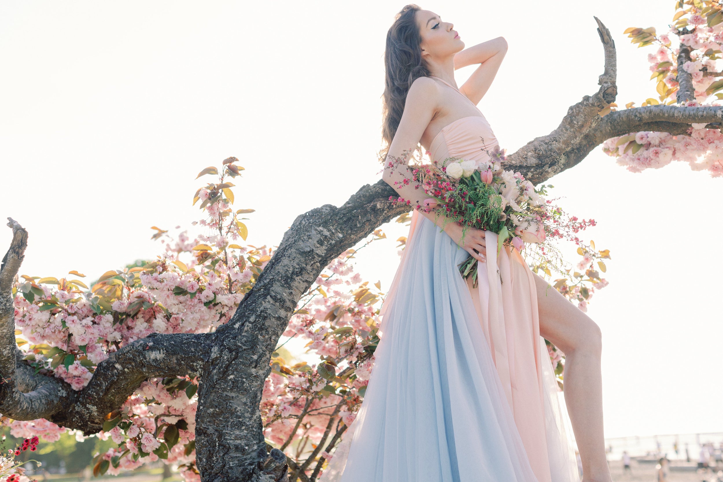 A woman posing outdoors on a flowering tree branch, wearing a strapless pastel gown with a pink bodice and a flowing light blue skirt, holding a bouquet of pink and white flowers.