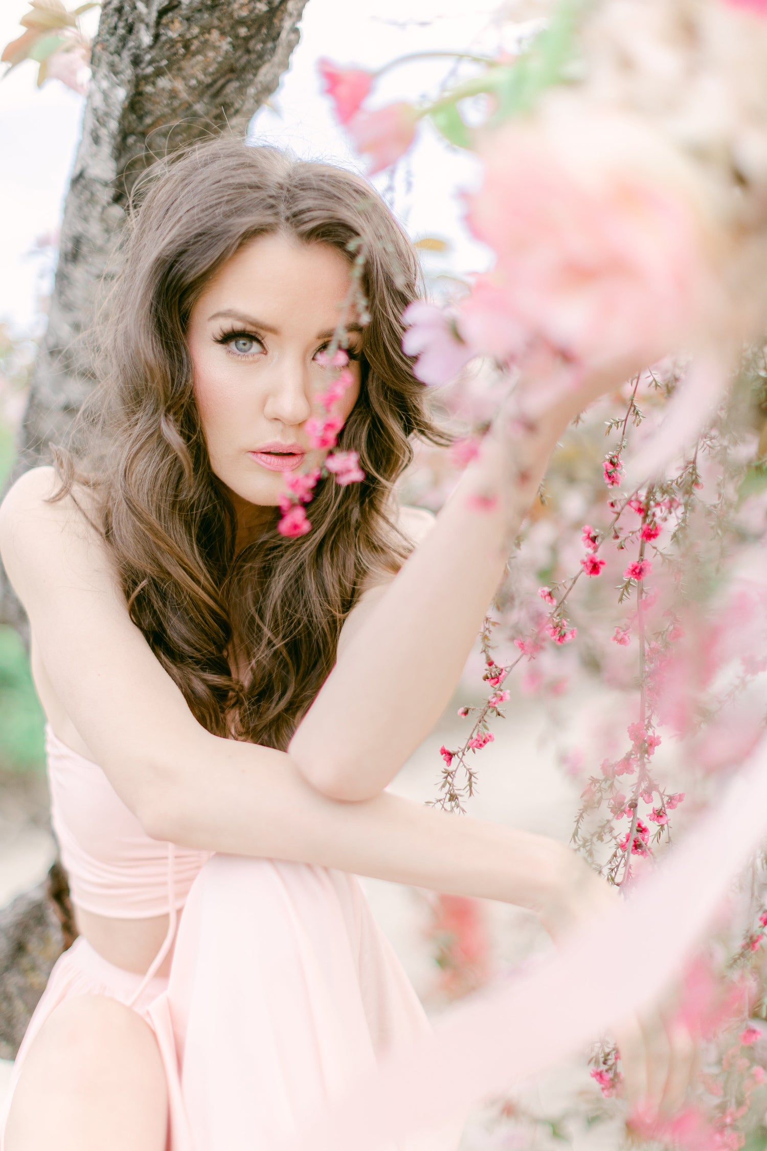 A woman with long wavy brown hair wearing a light pink sleeveless dress, posing outdoors next to a tree trunk with pink blossoms in the foreground.