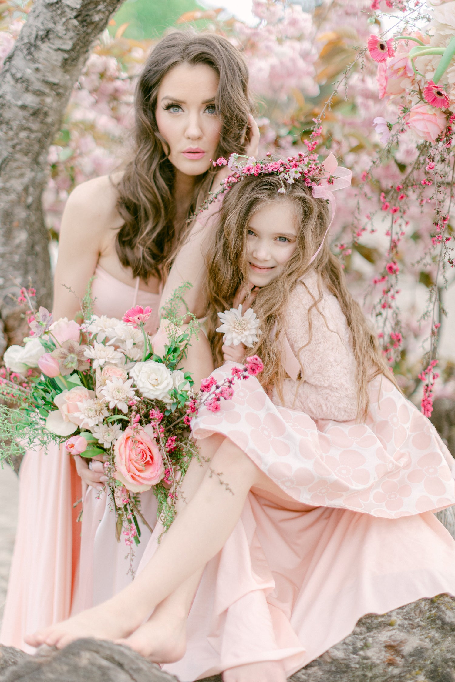 A woman and a young girl sit together outdoors among pink blossoms. The woman wears a pale pink strapless gown and holds a large bouquet of pink, white, and peach flowers.