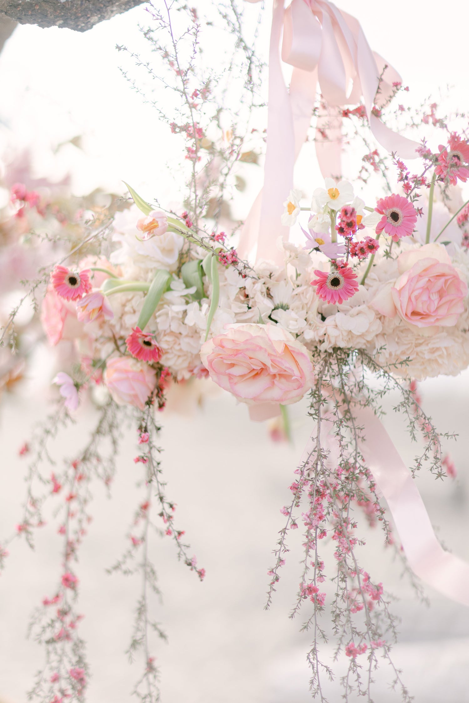 A soft-focus floral arrangement in pastel pink, white, and blush tones with delicate ribbons, photographed against a bright white background.