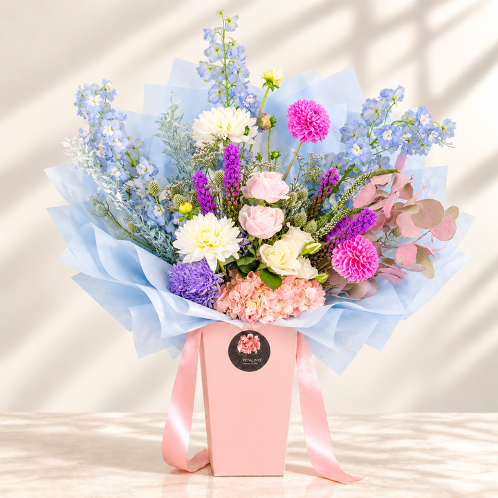 A vibrant floral bouquet in a pink box with a light blue wrapper, featuring pink, purple, white, and blue flowers, set against a warm beige background with soft window shadow patterns.