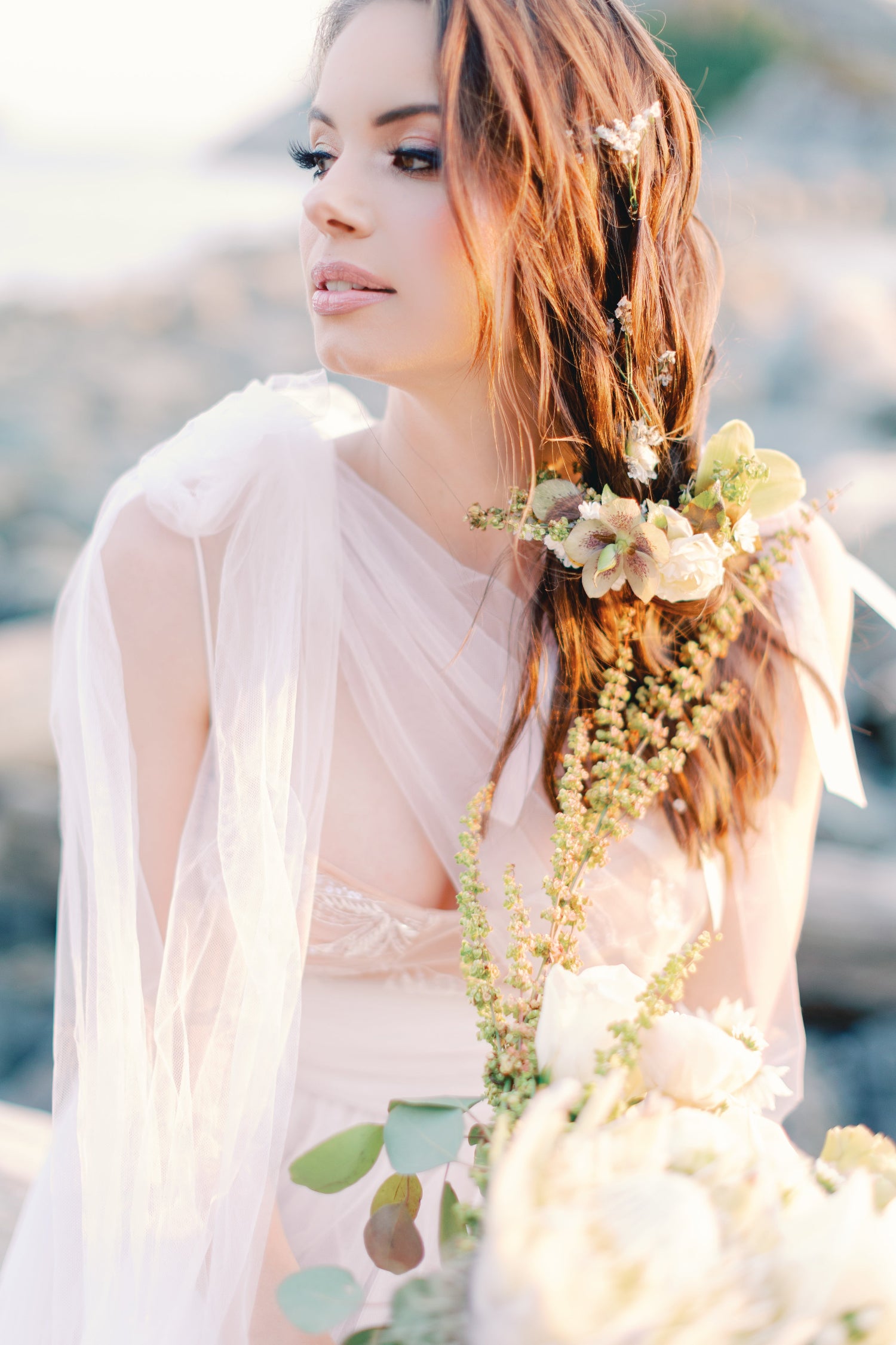 A woman outdoors wearing a sheer white bridal veil and a light blush pink lace wedding dress, holding a bouquet of white and cream flowers with greenery. Her hair is styled with a floral crown, and the background is a softly blurred natural outdoor setting.