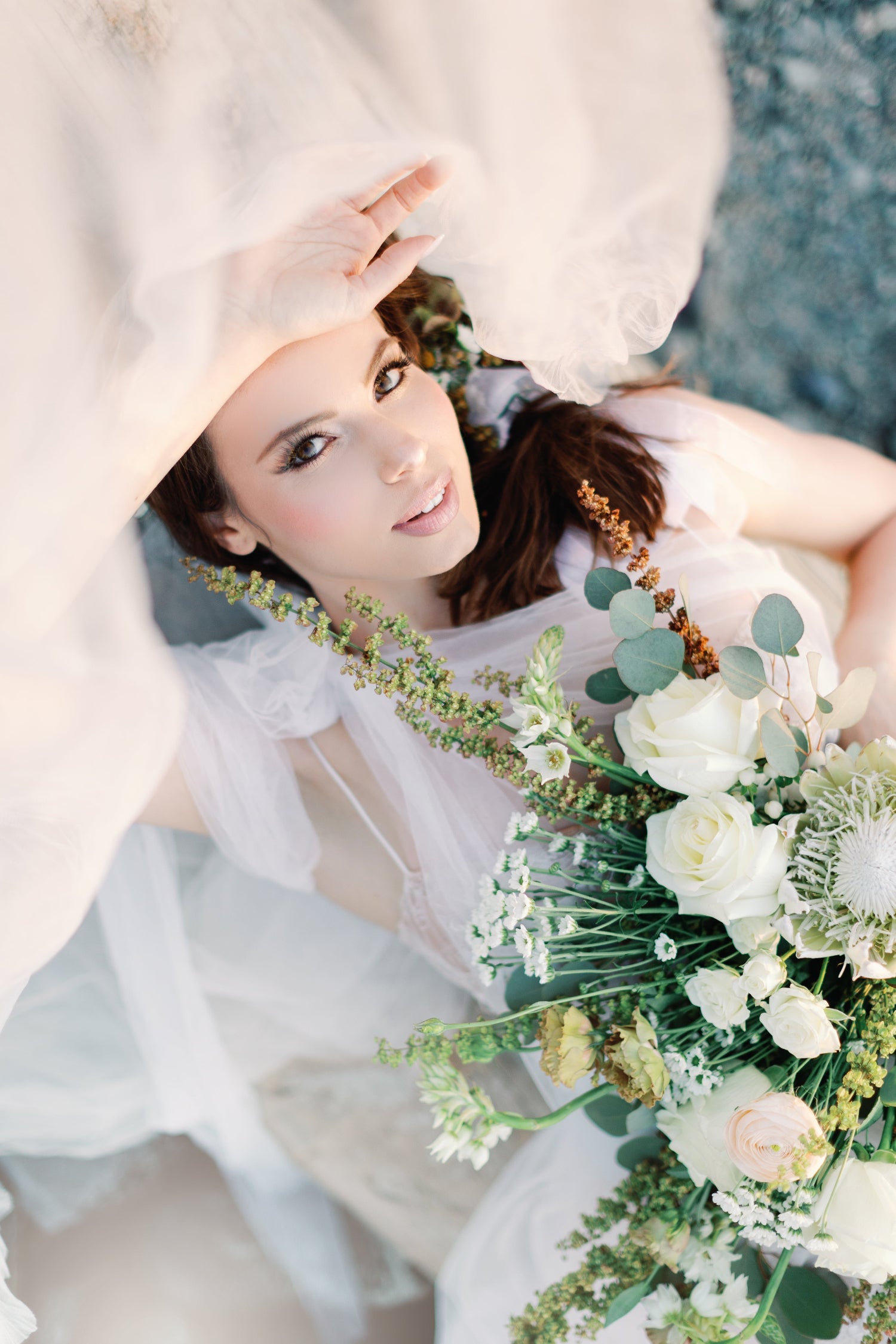 A woman in a white bridal gown and veil lies on a blue textured surface, holding a large bouquet of white and pale pink flowers with green foliage.