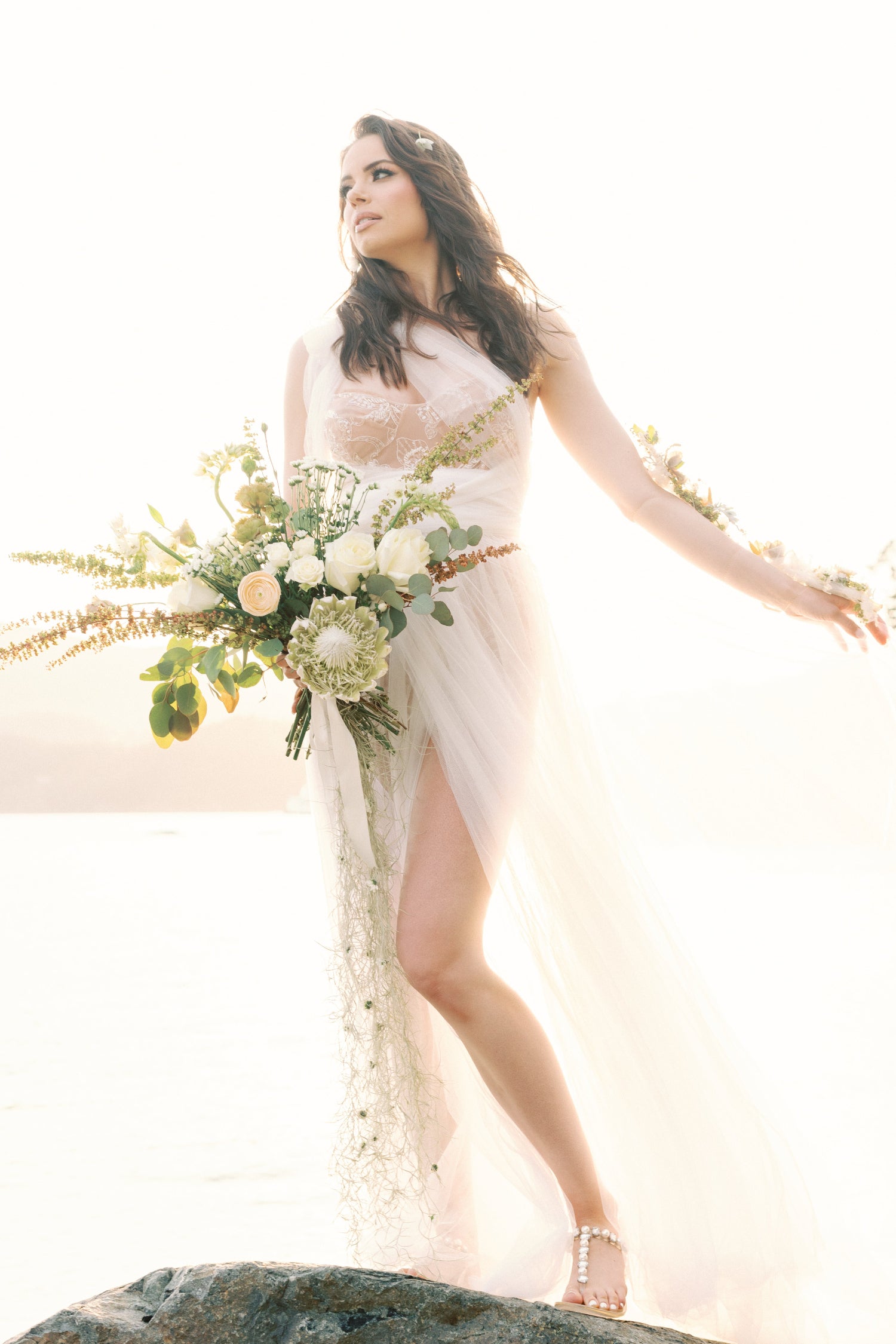 A woman in a flowing white wedding gown with a thigh-high slit stands on a rock by the sea, holding a large floral bouquet. The background is bright, overexposed white with soft sunlight and haze.