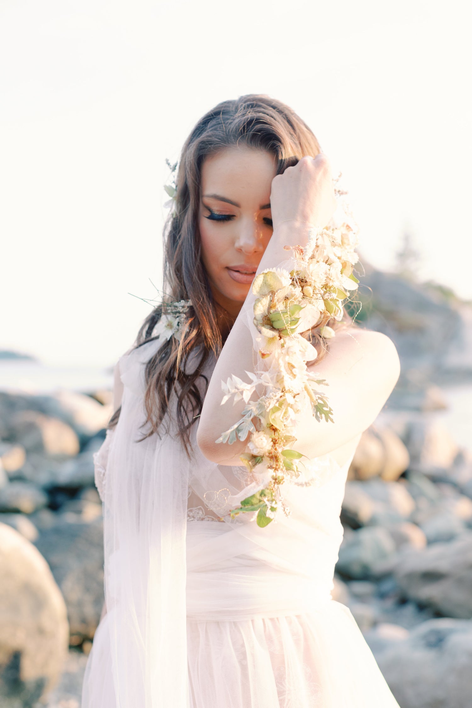 A woman in a flowing white dress stands on a rocky beach, holding a floral garland around her arm. The background is softly blurred with rocks and bright sunlight.