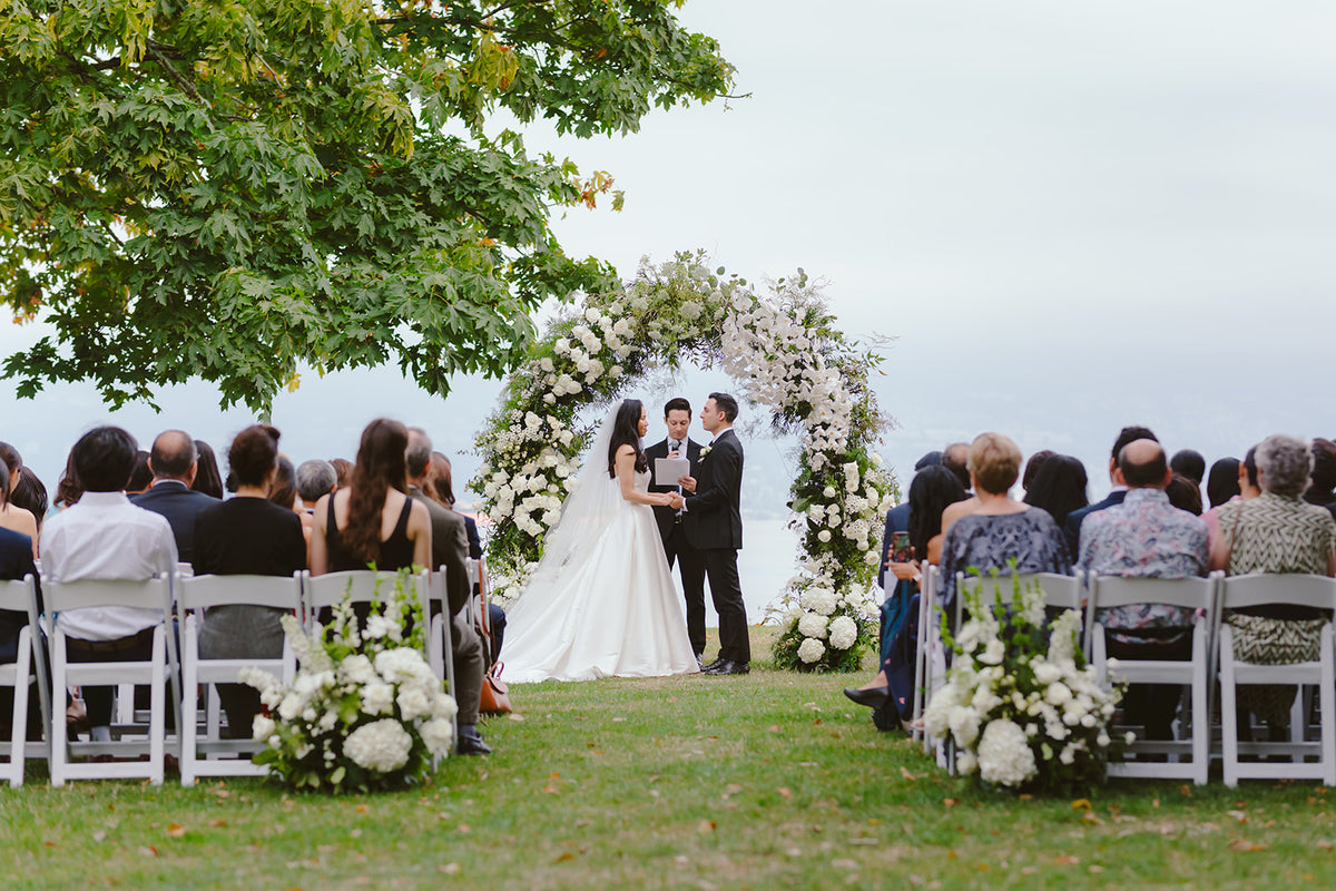 Outdoor wedding ceremony with a bride and groom standing under a floral arch, seated guests on white chairs on both sides, and a grassy lawn with trees in the background.