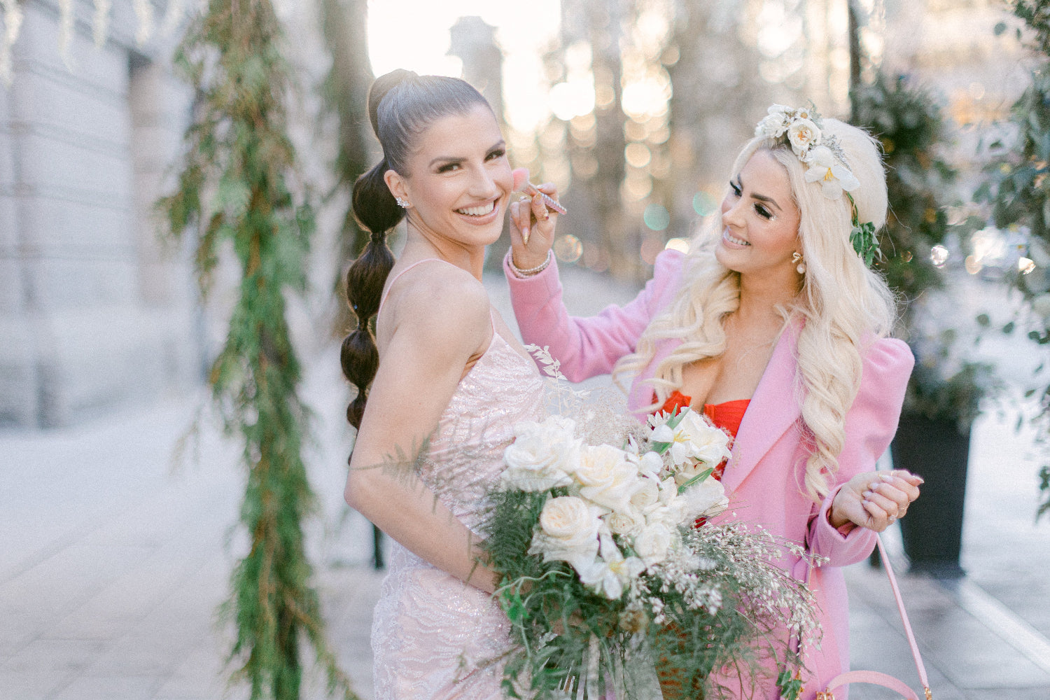 Two women outdoors in a softly blurred urban setting. One wears a pale pink sleeveless dress and holds a bouquet of white flowers, while the other wears a bright pink blazer over a red dress and has a floral headpiece.