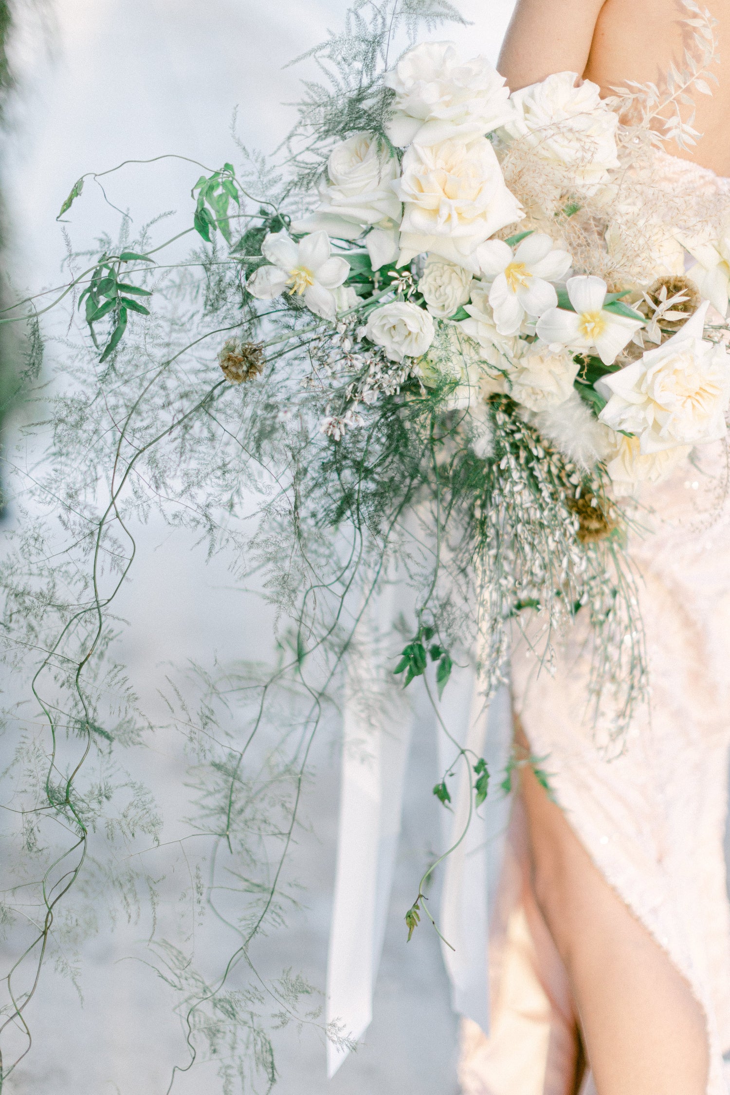 A close-up of a bridal bouquet with white roses, green foliage, and delicate ribbons, held by a bride wearing a pale blush lace dress.