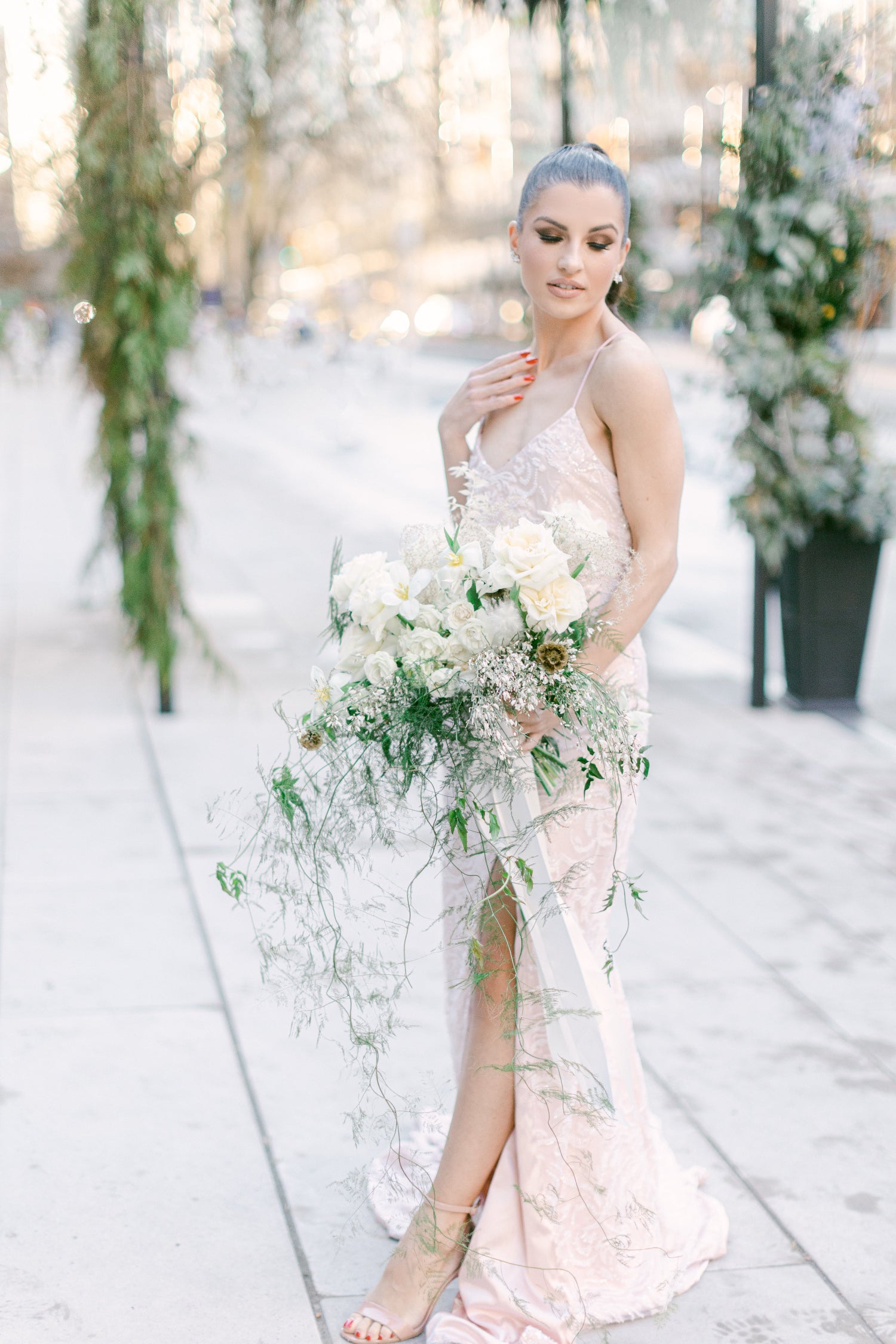 A woman in a pale blush pink sleeveless gown holding a large white and green floral bouquet, standing outdoors on a sunlit walkway with blurred greenery and planters in the background.