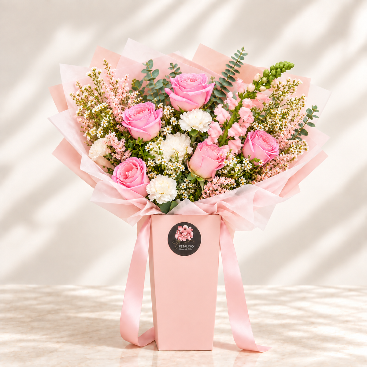 A pink floral bouquet in a pink gift box with a pink ribbon, featuring pink roses, white carnations, baby's breath, and green foliage, photographed on a light surface with soft shadow patterns.