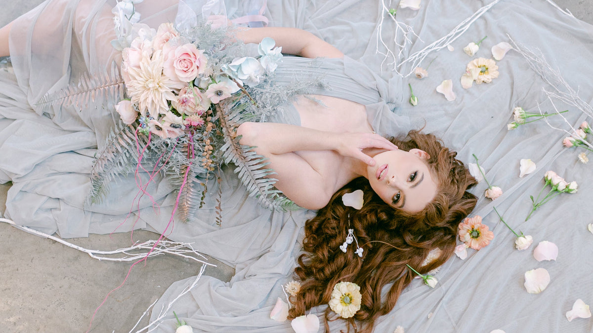 A woman with long wavy brown hair lies on a gray concrete floor surrounded by sheer light gray fabric, scattered flower petals, and a pastel bouquet. She is wearing a soft sheer gray dress or gown.