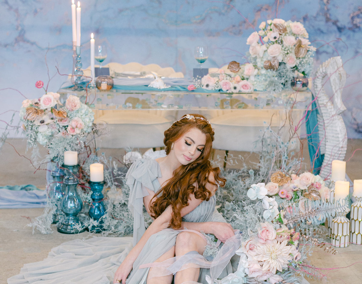 A woman in a flowing pale blue dress sits on the floor in a styled event setting with floral arrangements, candles, and a blue marble backdrop.