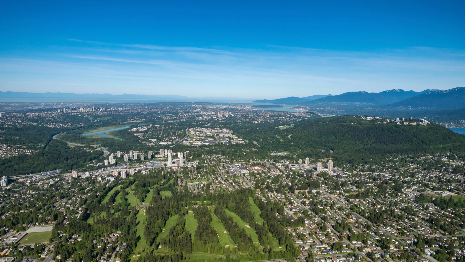 Aerial view of a dense suburban city with green golf courses, residential neighborhoods, a river bend, and distant mountains under a clear blue sky.