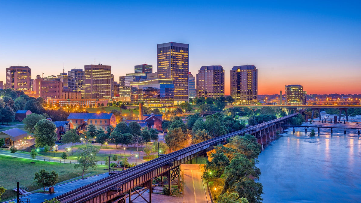 A scenic city skyline at dusk with illuminated skyscrapers, a river in the foreground, a bridge, and trees under a blue-to-orange sunset sky.