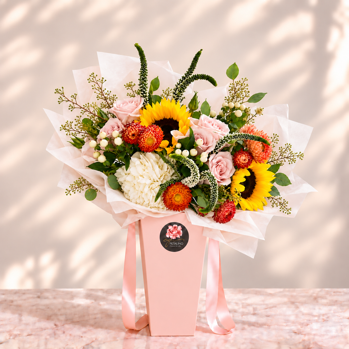 A vibrant mixed floral bouquet in a light pink gift box with pink satin ribbons, set against a warm beige background with soft leaf shadows. The bouquet features yellow sunflowers, pink roses, white chrysanthemums, and red-orange pom-pom flowers with green foliage.