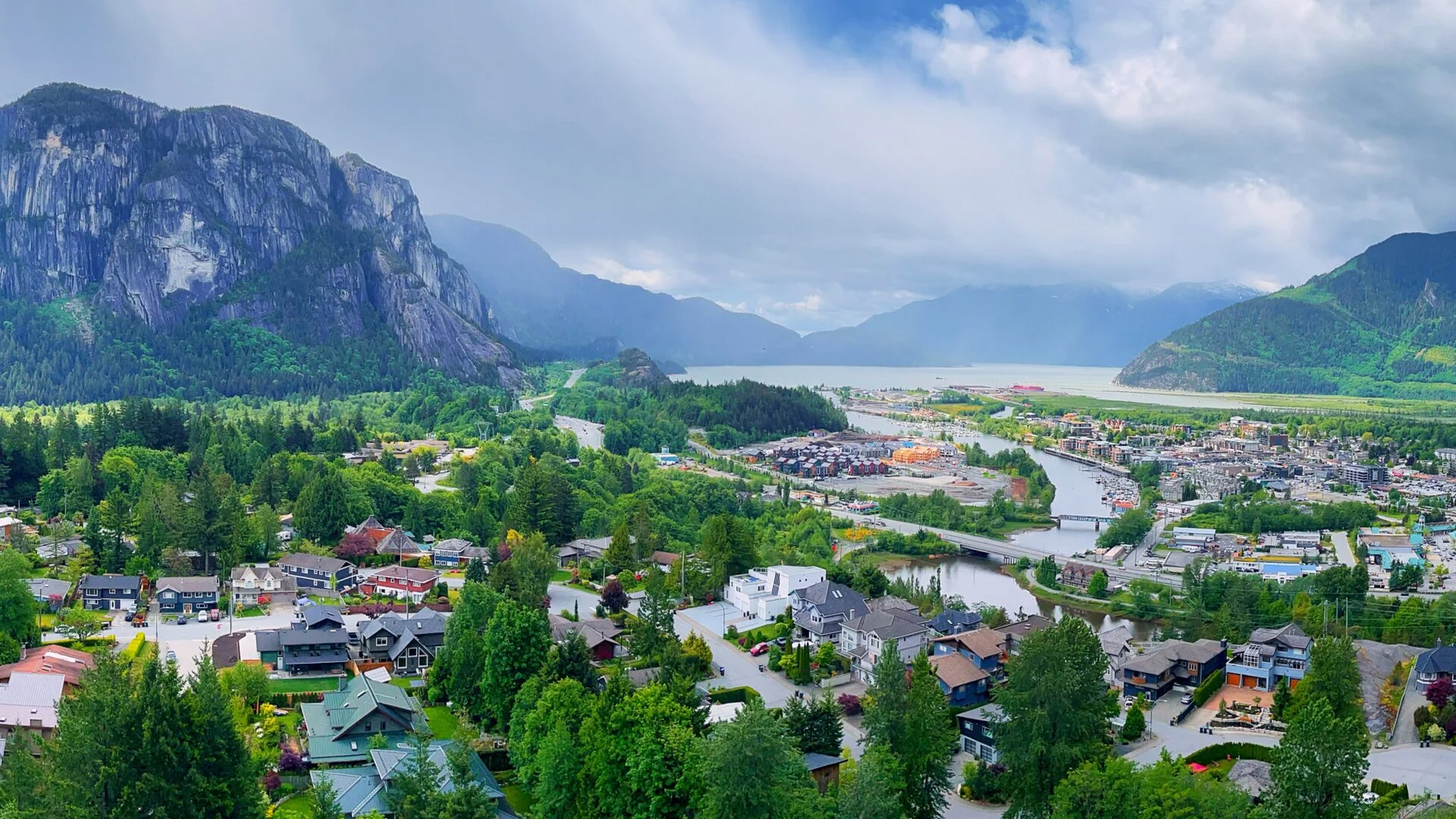 A scenic landscape photograph of a small town nestled in a valley between steep mountains, with a river flowing through the center, surrounded by green trees and a dramatic blue sky with white clouds.