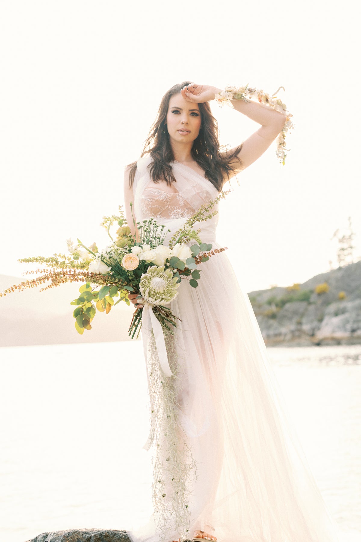 A bride in a sheer blush pink lace wedding gown stands on a beach holding a large floral bouquet, with a bright sunlit sky and distant shoreline in the background.