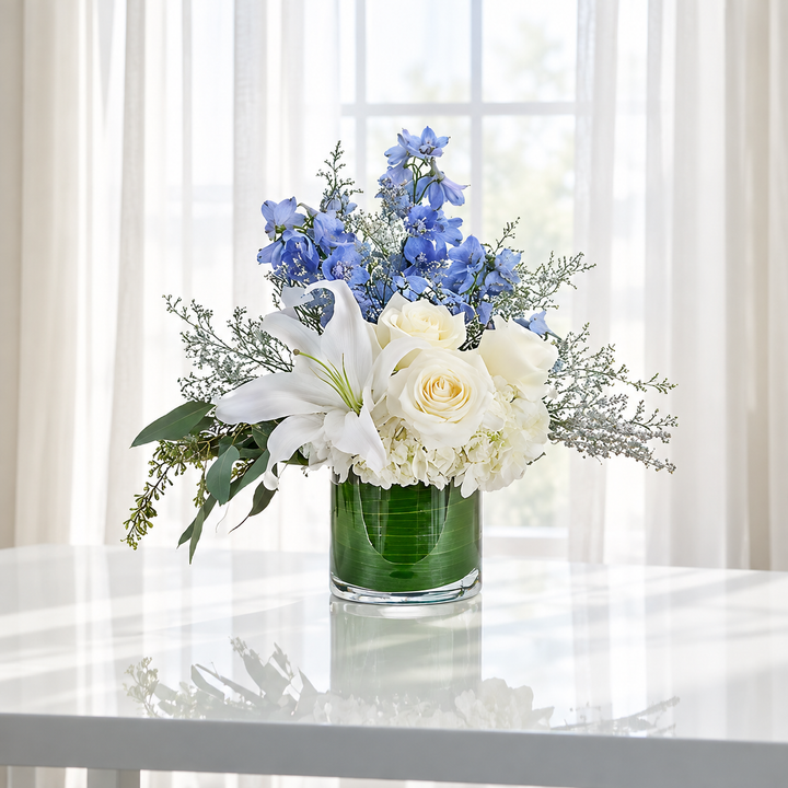 A floral arrangement in a clear glass vase with green water, featuring white lilies, cream roses, blue flowers, and green foliage, displayed on a white table in front of a bright window with sheer curtains.