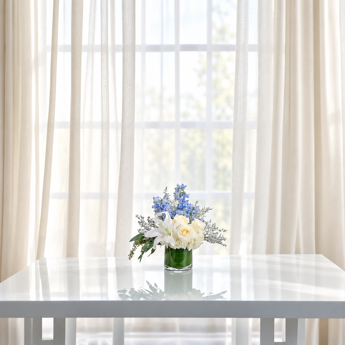 A small floral arrangement in a clear glass vase sits on a white table in front of sheer cream curtains and a bright window.