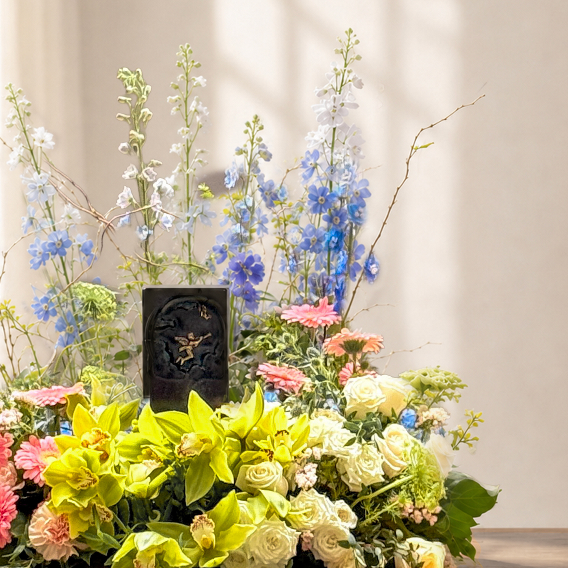 A floral arrangement on a wooden table featuring yellow lilies, white roses, pink daisies, blue delphiniums, and a small black urn centerpiece, styled in a bright room with a window and a turquoise butterfly candle holder beside it.