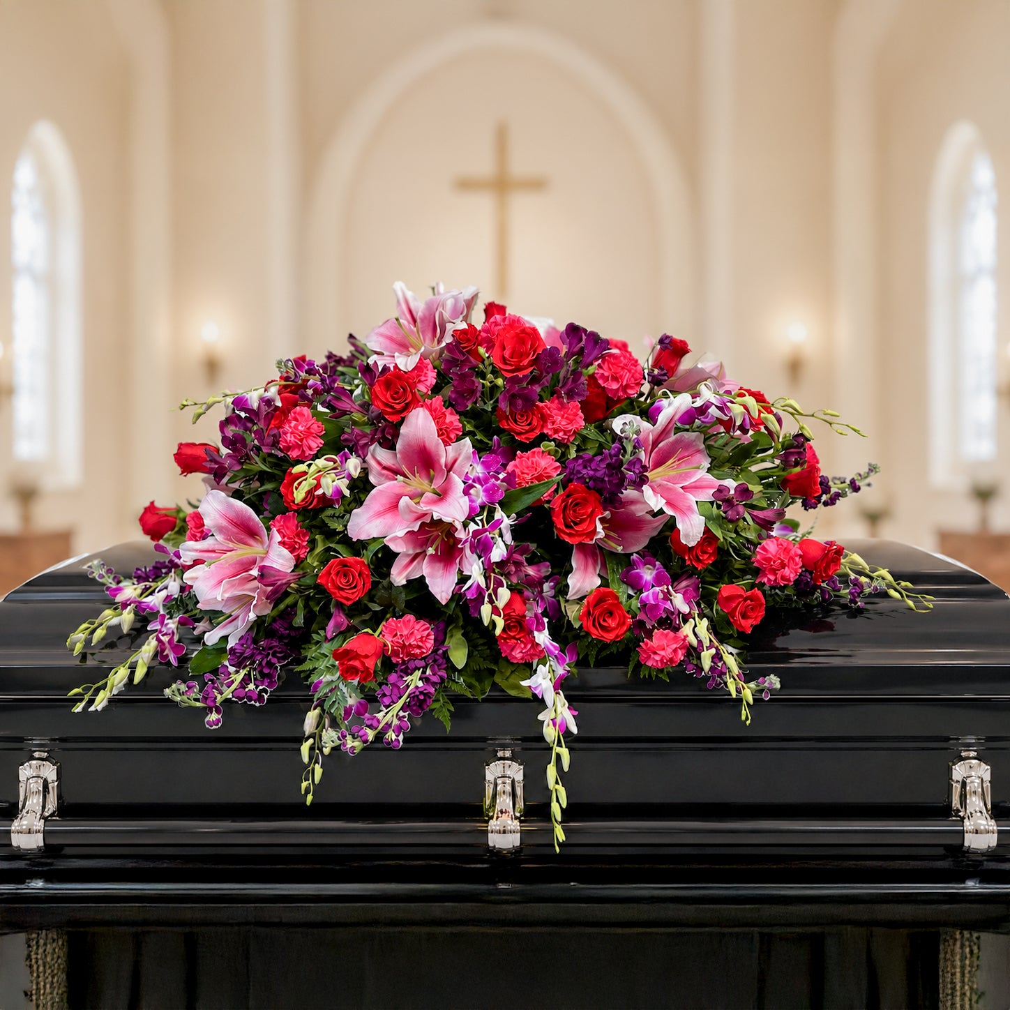 A large, vibrant floral arrangement featuring pink lilies, red roses, and purple orchids, displayed on a black casket in a softly blurred church interior.