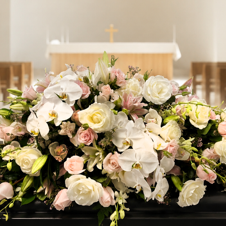 A floral arrangement of white orchids, pink roses, and cream lilies with green foliage draped over a black casket in a church setting.