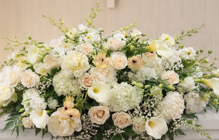 A white casket in a chapel setting, decorated with a large floral arrangement of white, cream, and pale yellow roses with green foliage. A wooden cross is visible on the arched wall behind the casket.