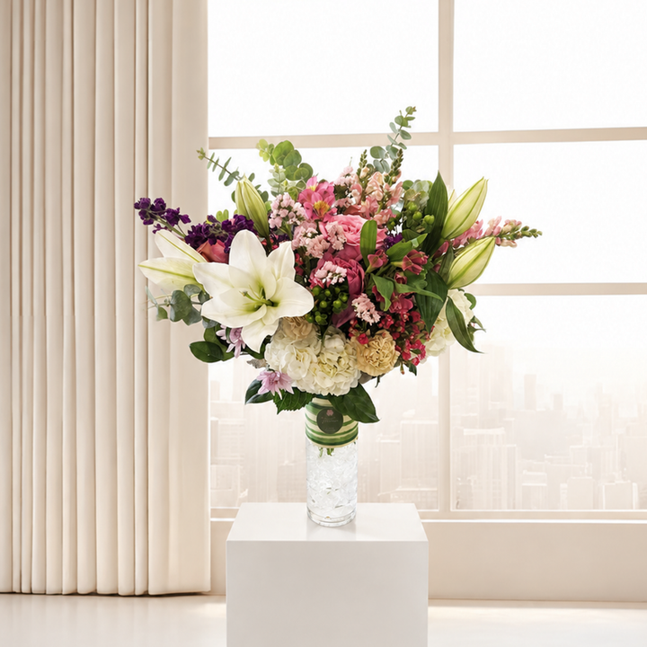 A vibrant mixed floral arrangement featuring a large white lily, pink roses, purple stock, cream hydrangeas, and green foliage, presented in a striped vase against a soft, light beige indoor background.