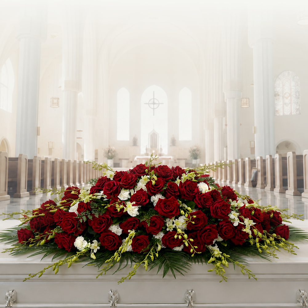 A large floral arrangement of red and white roses with green foliage draped over a white casket in a bright, softly lit church interior with pews and tall columns.