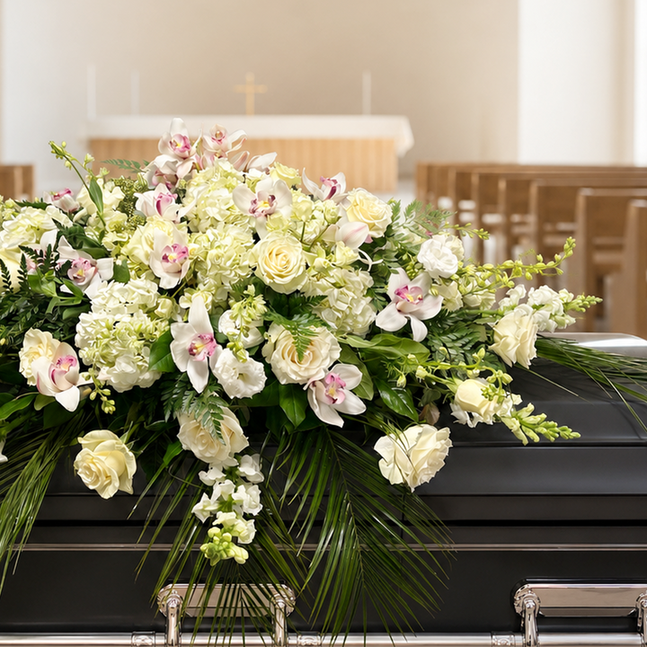 A large floral casket arrangement with white and pale pink flowers and green foliage displayed on a black casket in a church interior.
