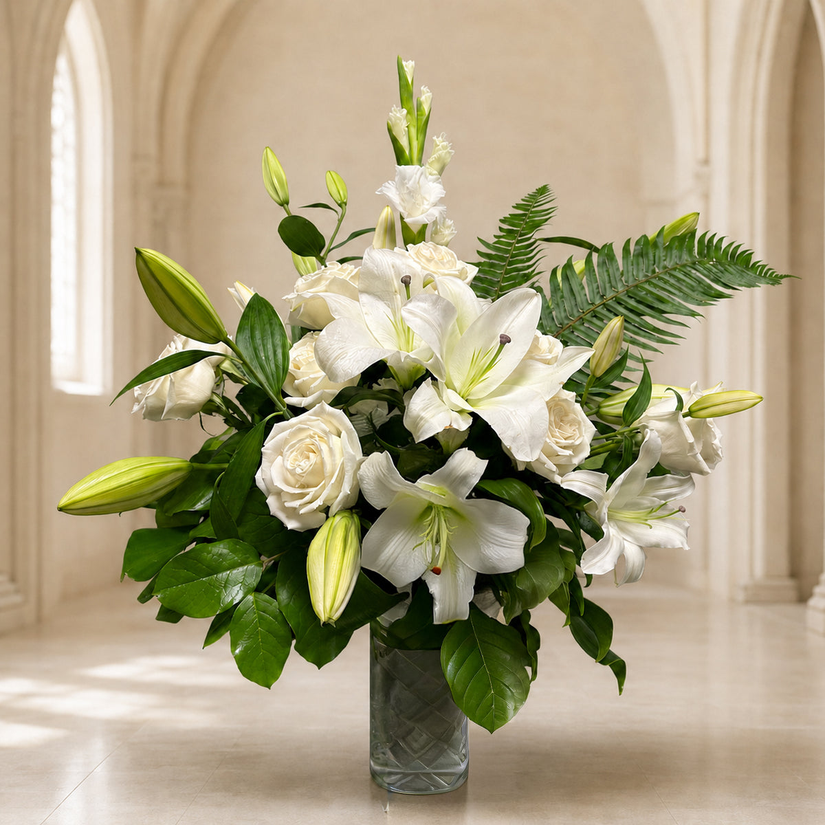 A sympathy flower arrangement featuring white lilies, white roses, and green foliage in a clear glass vase, 