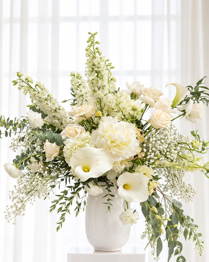 A large white ceramic vase filled with a lush floral arrangement of white and cream flowers and green foliage, displayed on a white pedestal in a bright, sunlit room with sheer white curtains.