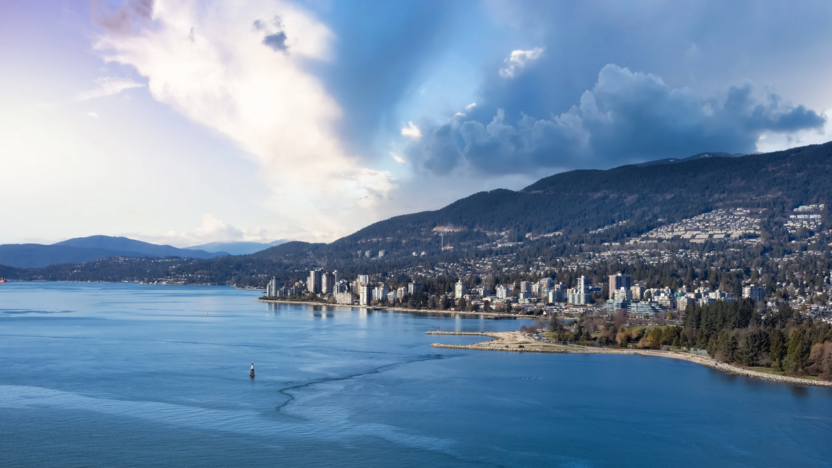 Panoramic view of a coastal city with high-rise buildings along a bay, surrounded by mountains and dramatic clouds, with blue water in the foreground.