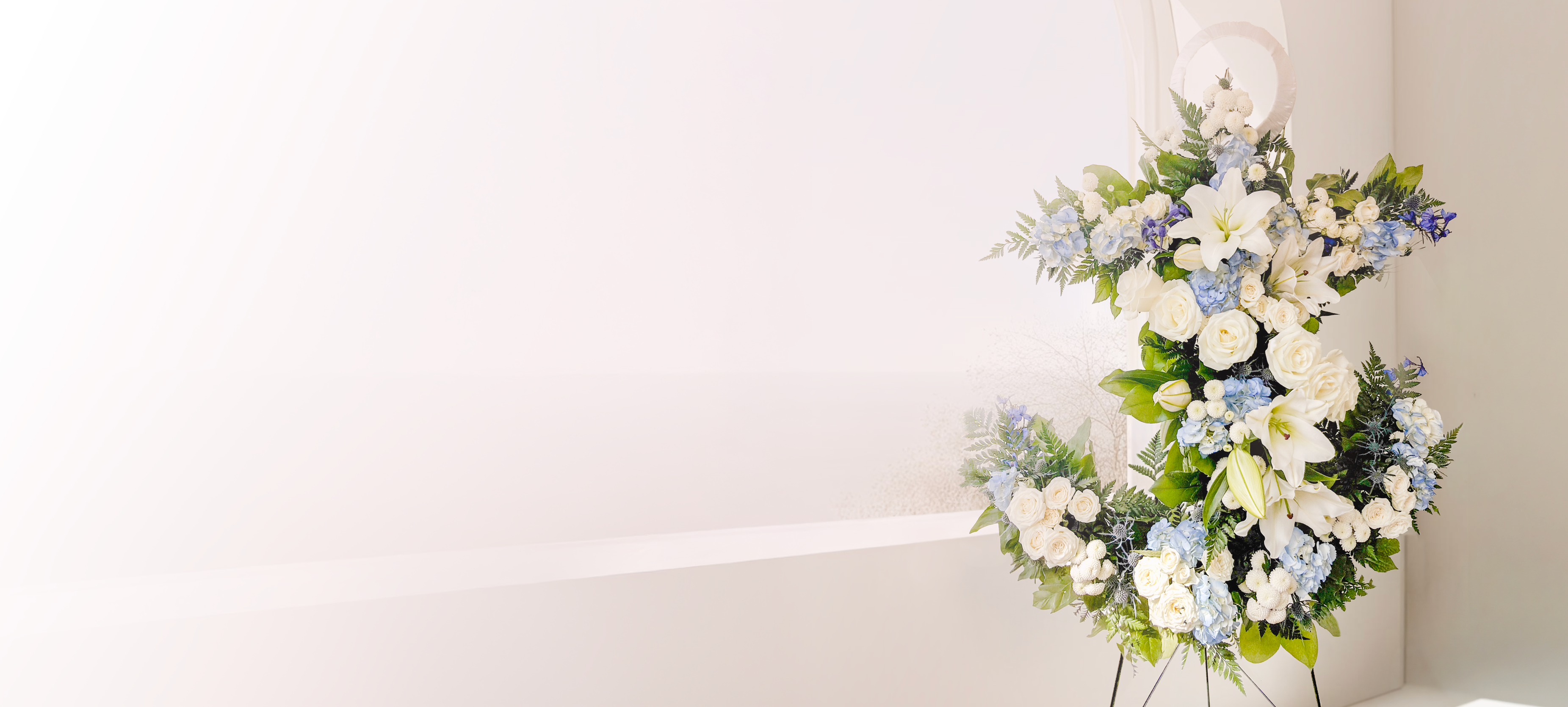 A large floral arrangement featuring white and light blue flowers with green foliage, displayed on a stand against a clean white wall with a shelf.