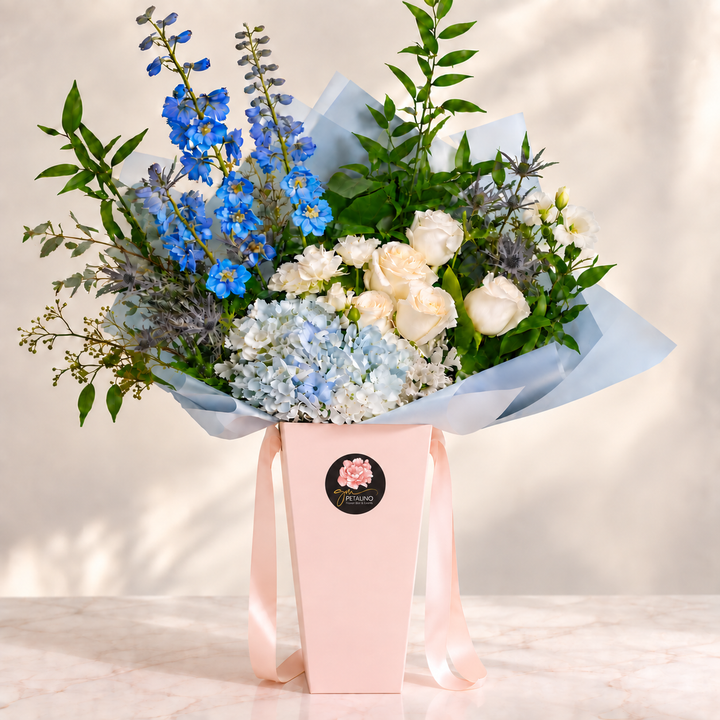 A floral bouquet arranged in a pale pink box with a light pink ribbon and a round black logo sticker. The bouquet includes blue delphiniums, light blue hydrangeas, white roses, green foliage, and soft lavender accents, set against a softly lit neutral background.