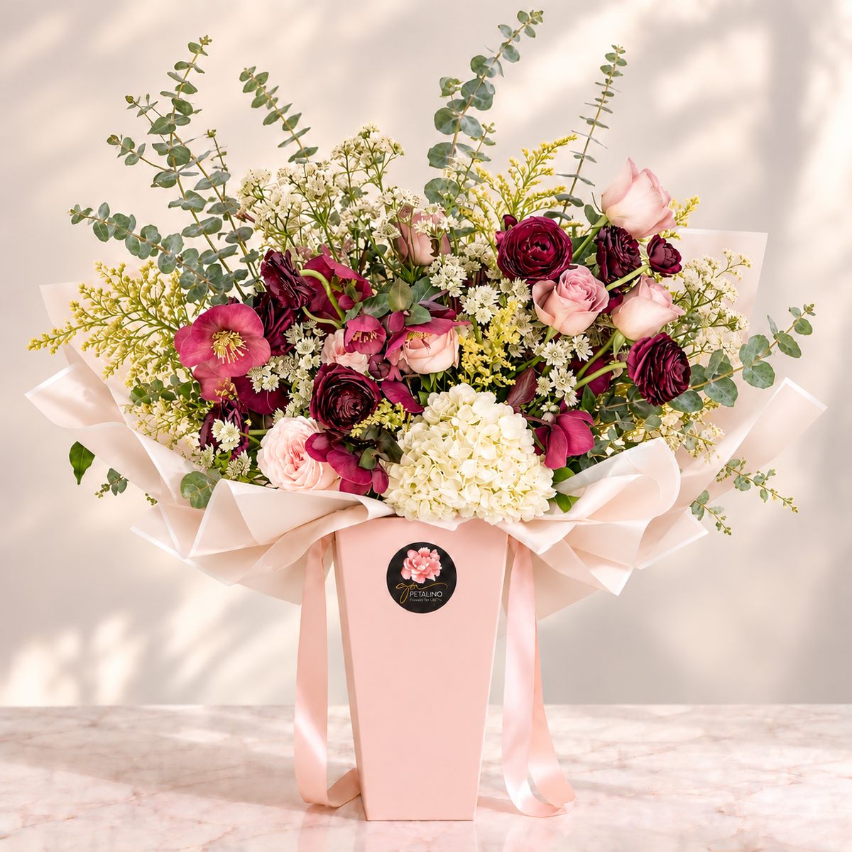 A floral arrangement in a pale pink box with a pink ribbon and a round black logo sticker. The bouquet contains burgundy and pink roses, light pink roses, white baby's breath, green eucalyptus stems, and white hydrangea-like blooms, set on a light marble surface against a softly lit neutral background.
