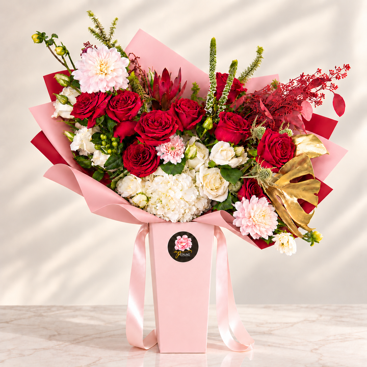 A large floral bouquet arranged in a pink gift box with pink wrapping paper and a gold ribbon bow, photographed on a light marble surface against a softly lit neutral background.