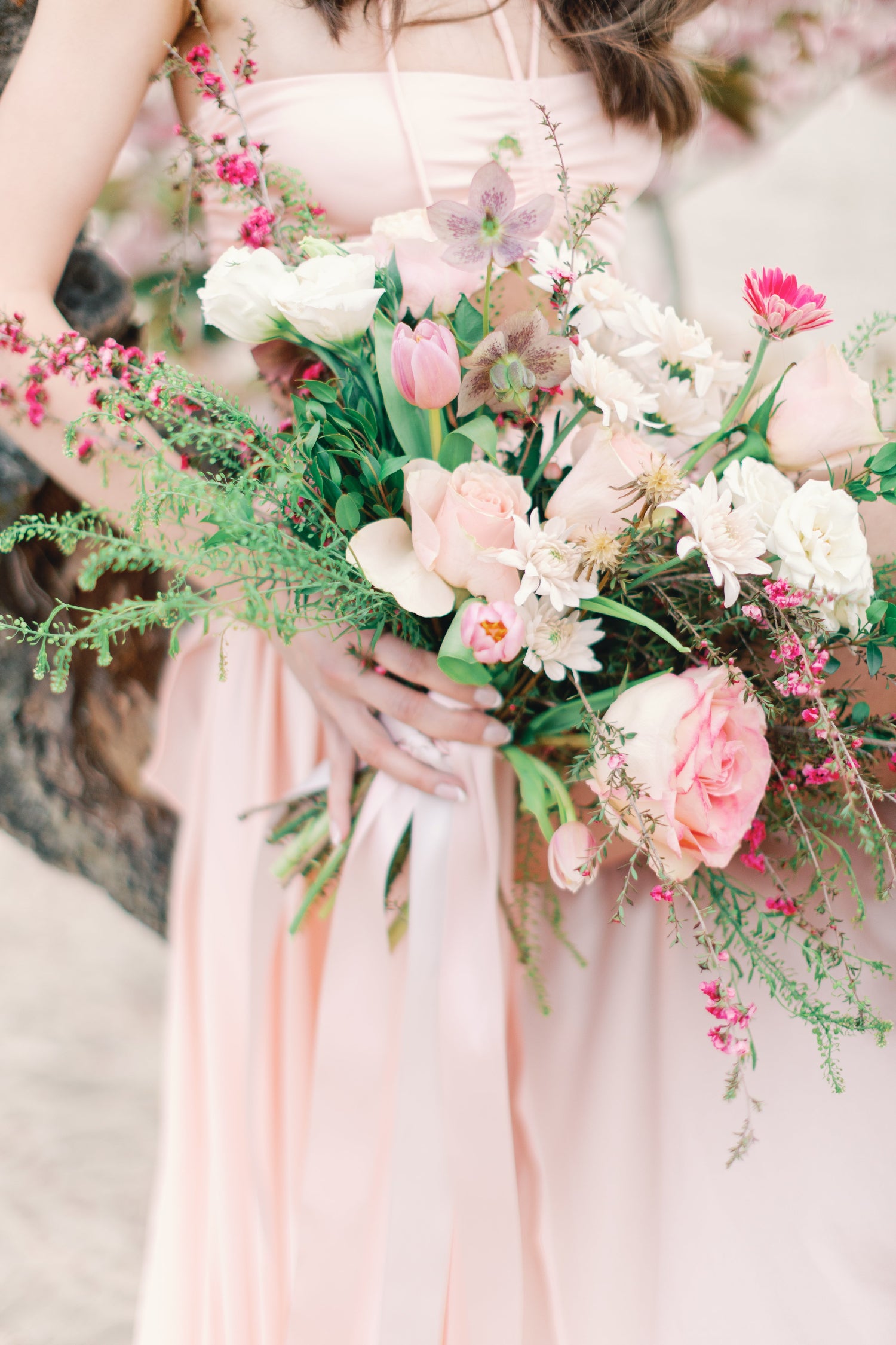 A close-up of a person in a soft blush pink dress holding a large floral bouquet with pink, white, and green flowers.