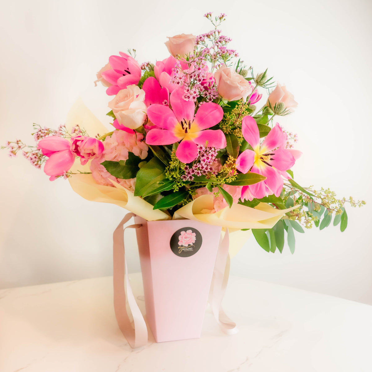 A vibrant flower bouquet featuring bright pink lilies, soft peach roses, and green foliage, presented in a light pink box with a ribbon and a circular floral logo sticker.