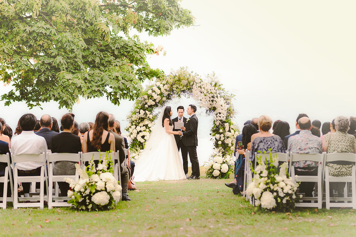 Outdoor wedding ceremony with a bride and groom standing under a floral arch, seated guests in white chairs on both sides, and green grass and trees in the background.