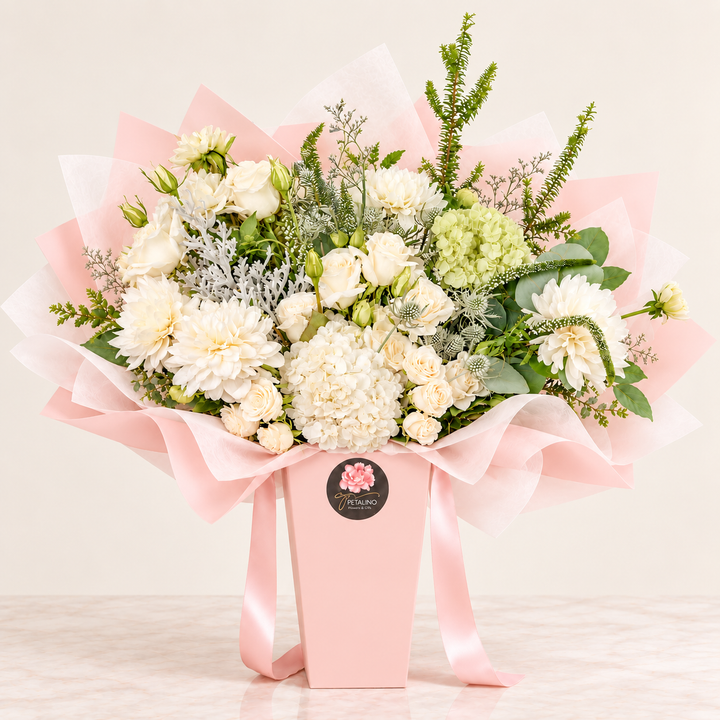 A floral bouquet arranged in a pink box with pink tissue paper and a pink ribbon, photographed on a light neutral surface against a pale beige background. The bouquet contains mostly cream and white flowers with green foliage and a few pale pink blooms.